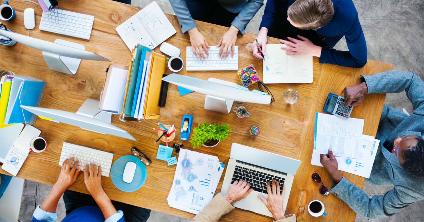 group of diverse people working at a large group desk, with coffee mugs and documents scattered about