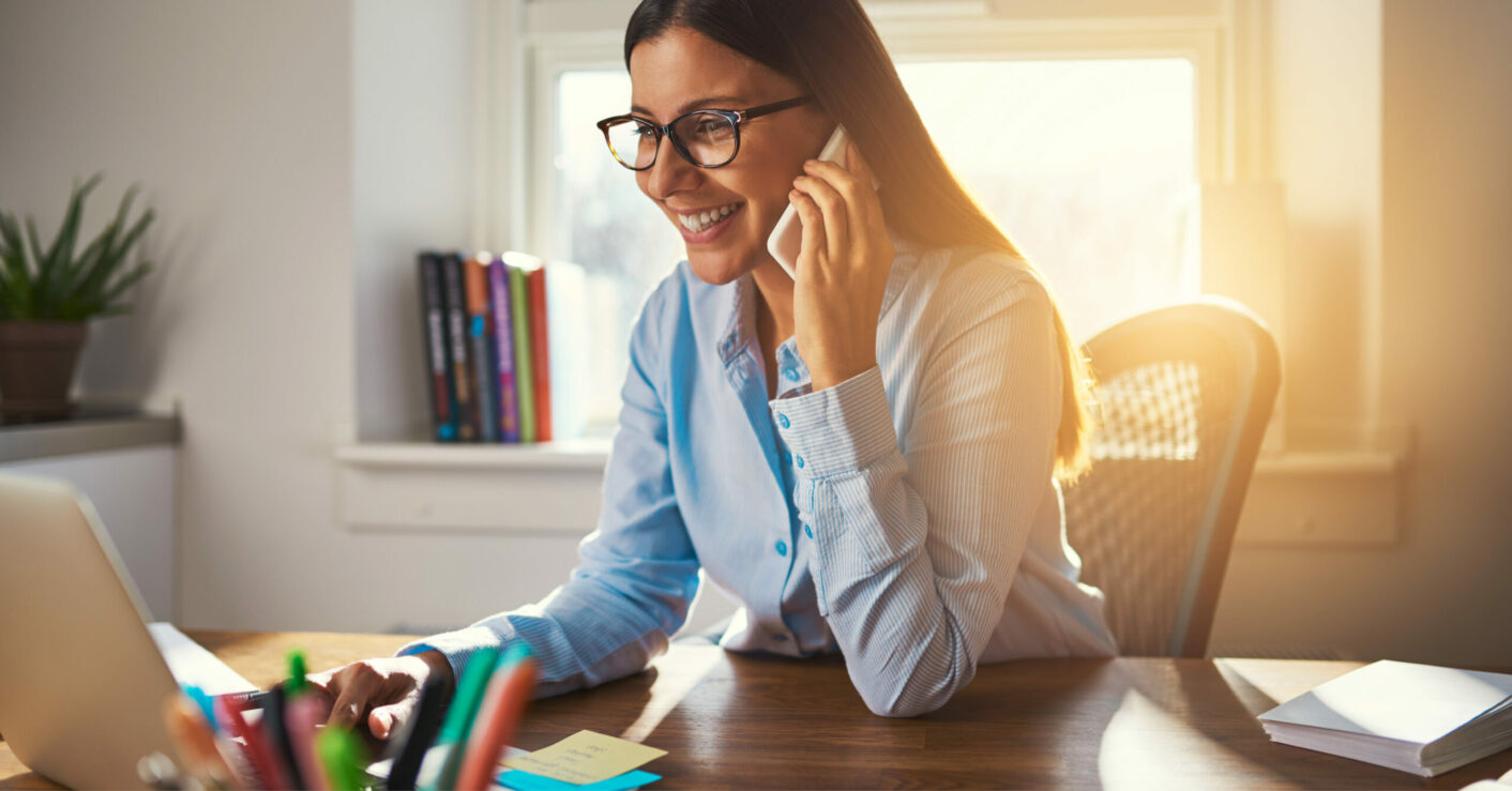 Smiling young woman at a desk in a sunny room, talking on a cell phone with one hand and working on a laptop with the other