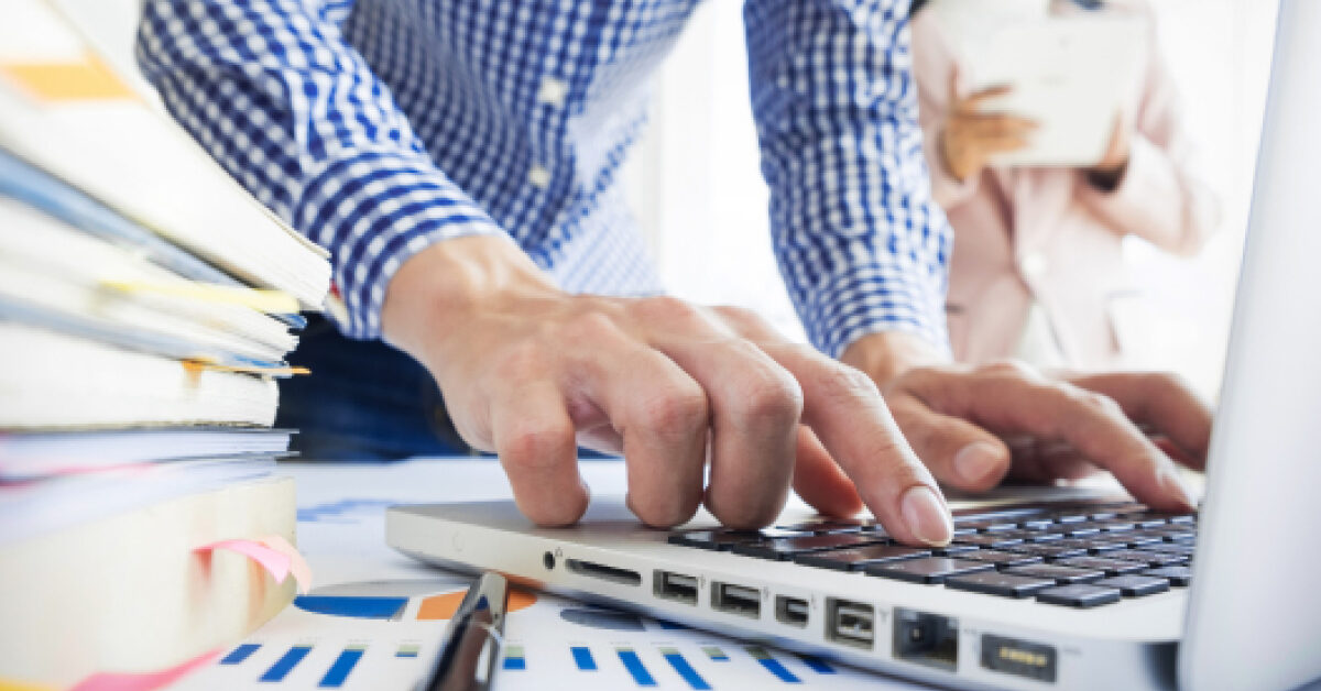 person busy typing on a laptop with important documents around on the desk